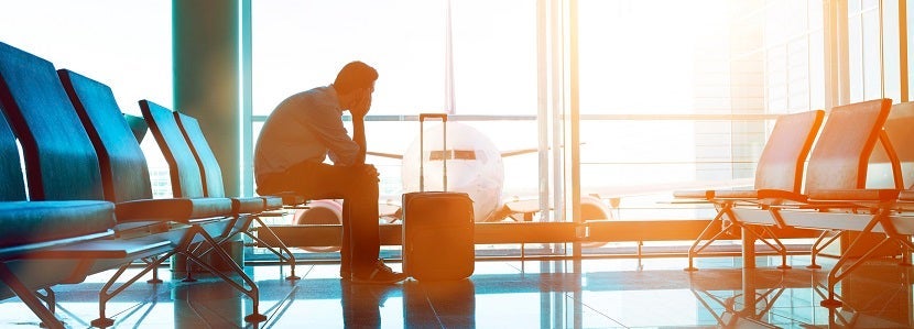 Man sits down in waiting area in airport terminal. He is ready for take off and looks out of the window at a plane. A suitcase stands before him.