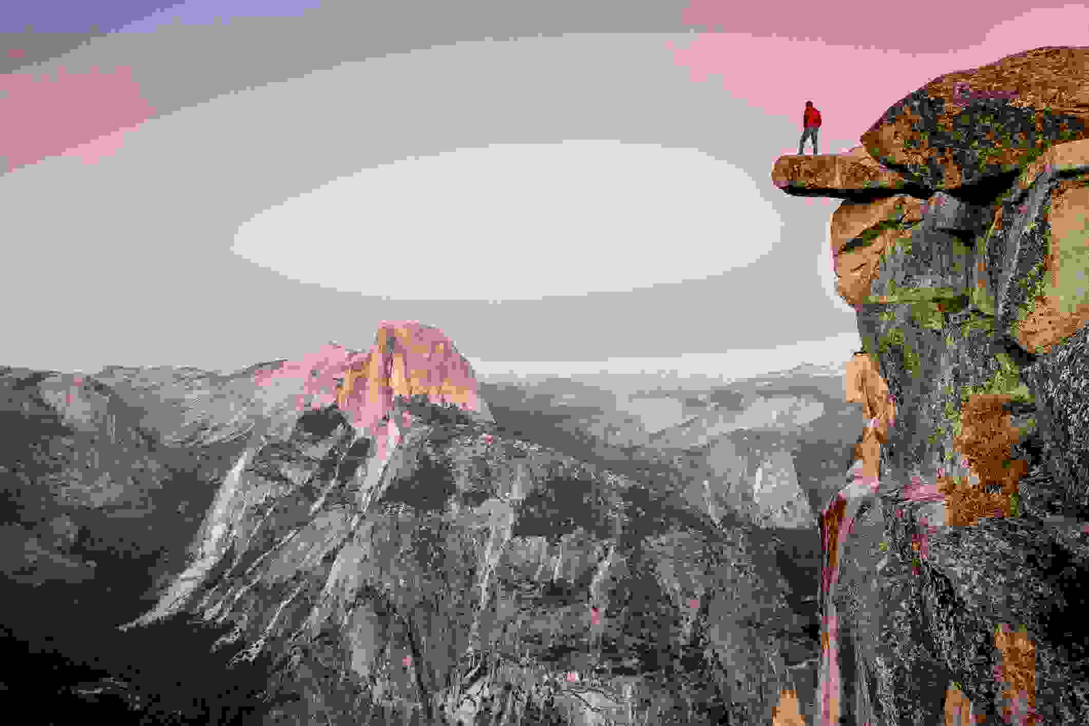 A fearless male hiker is standing on an overhanging rock at Glacier Point enjoying the breathtaking view towards famous Half Dome in beautiful post sunset twilight in summer, Yosemite National Park, California (Photo by bluejayphoto/Getty Images)