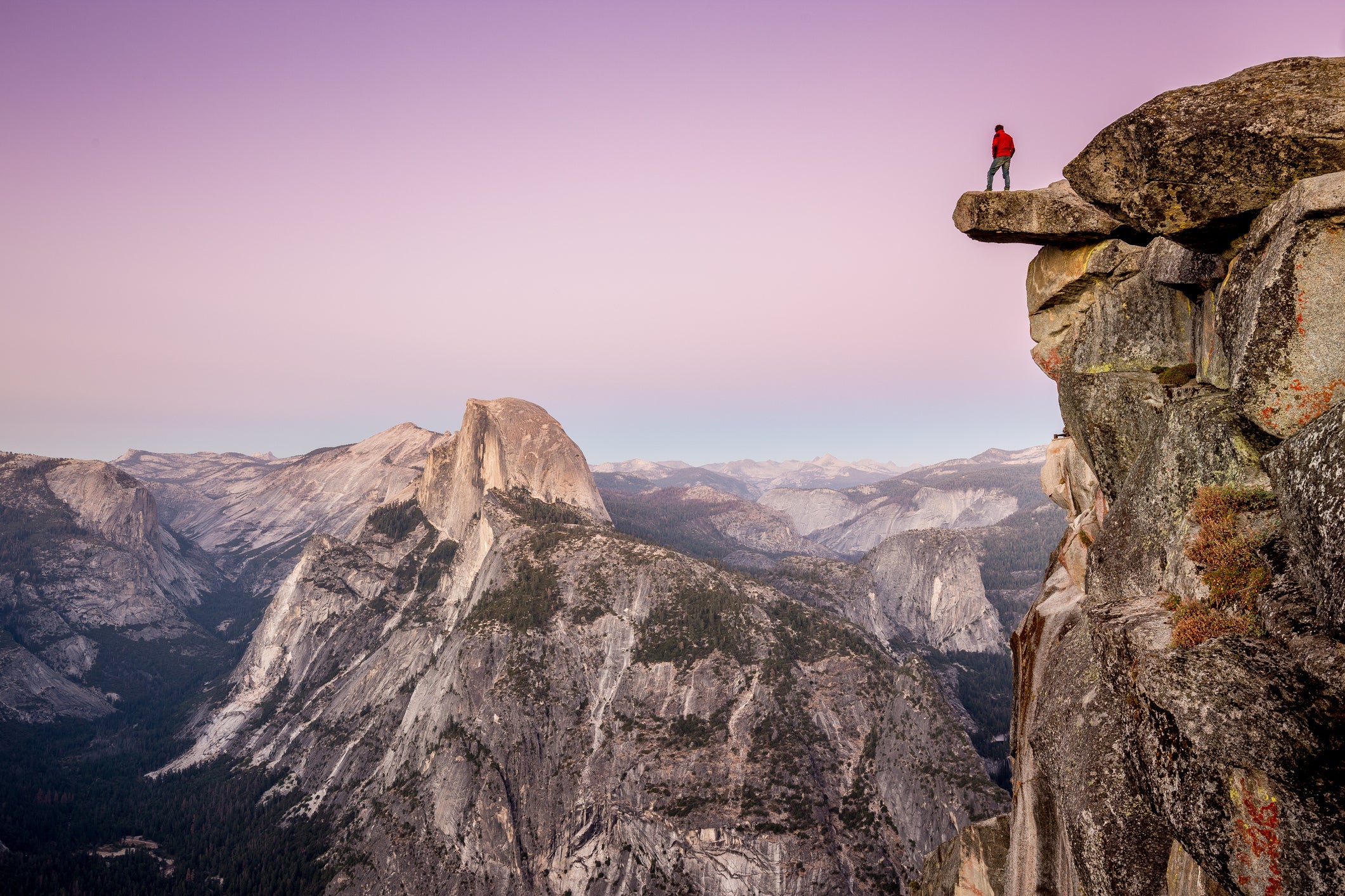 A fearless male hiker is standing on an overhanging rock at Glacier Point enjoying the breathtaking view towards famous Half Dome in beautiful post sunset twilight in summer, Yosemite National Park, California (Photo by bluejayphoto/Getty Images)