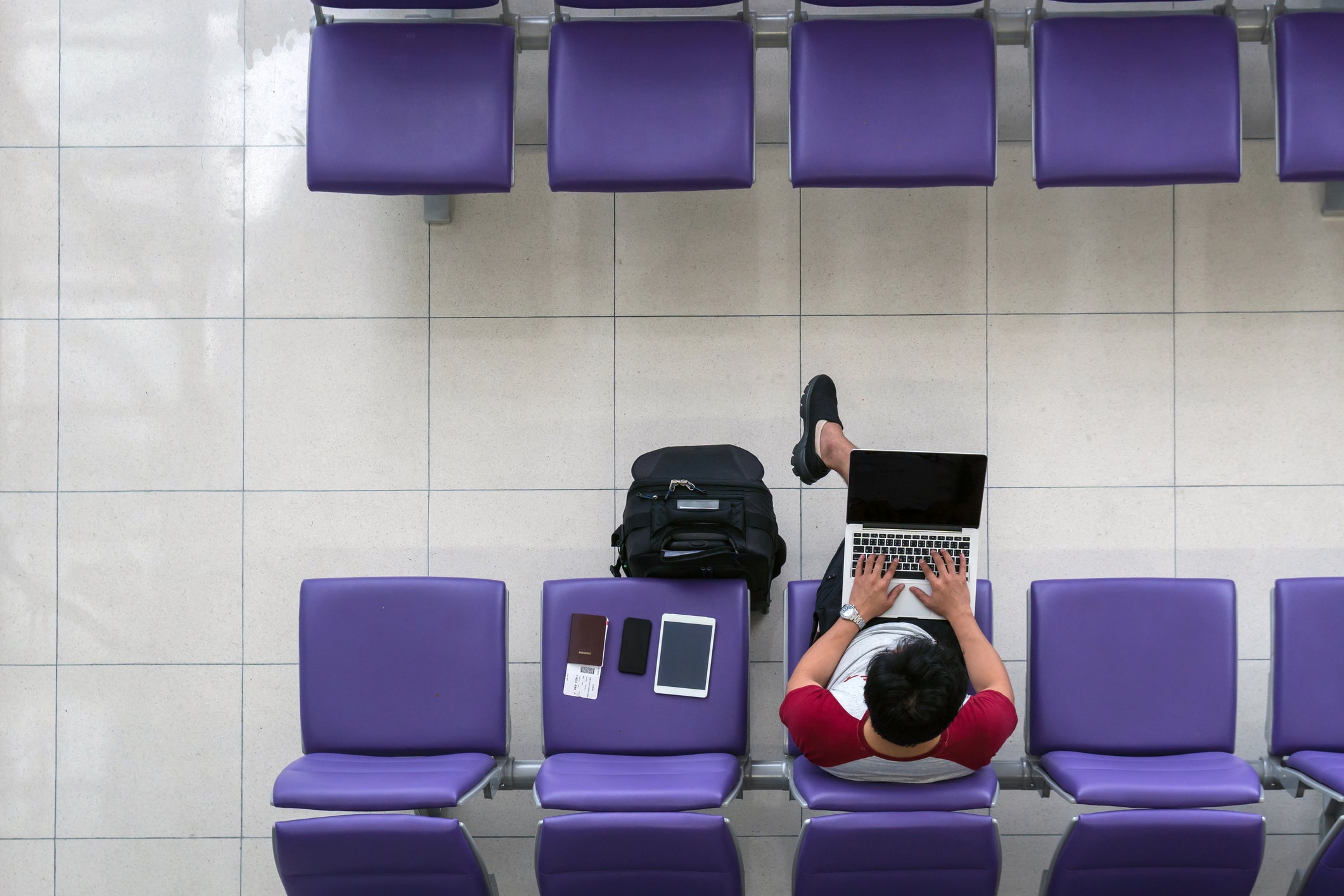 Happy Asian man traveler working with technology laptop with suitcase, tablet, smart mobile phone and passport at the seat while waiting airplane depart at the airport, freelanace and travel concept (Photo by Photographer is my life/GettyImages)
