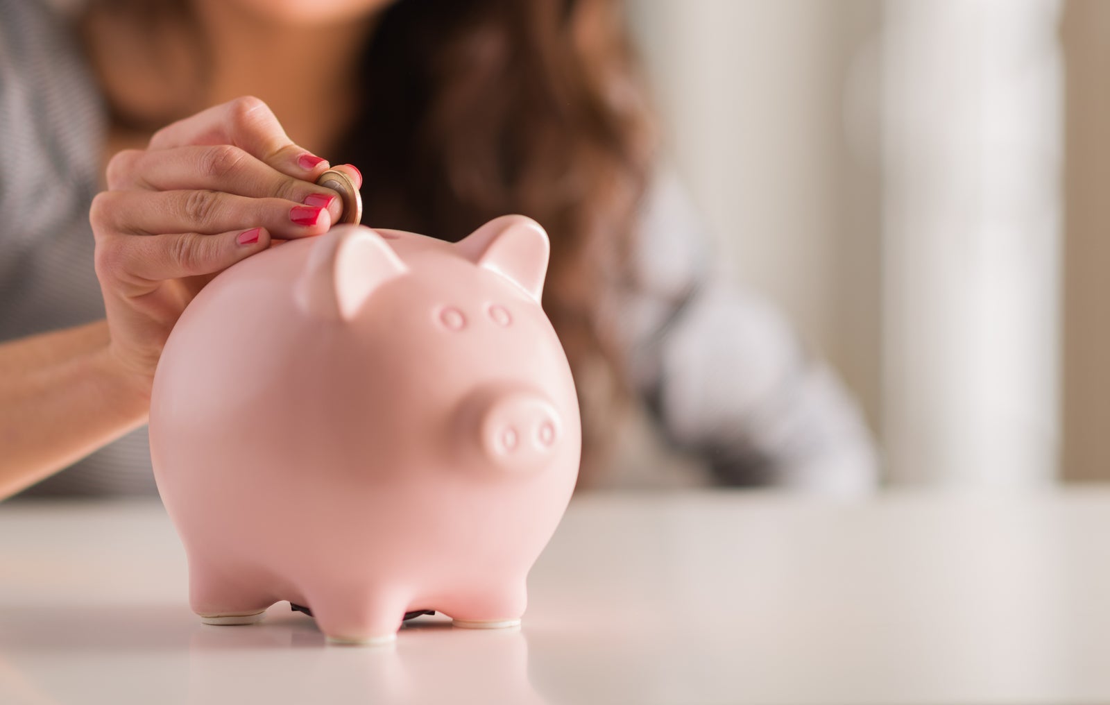 A woman with pink painted nails drops a coin into a pink piggy bank.