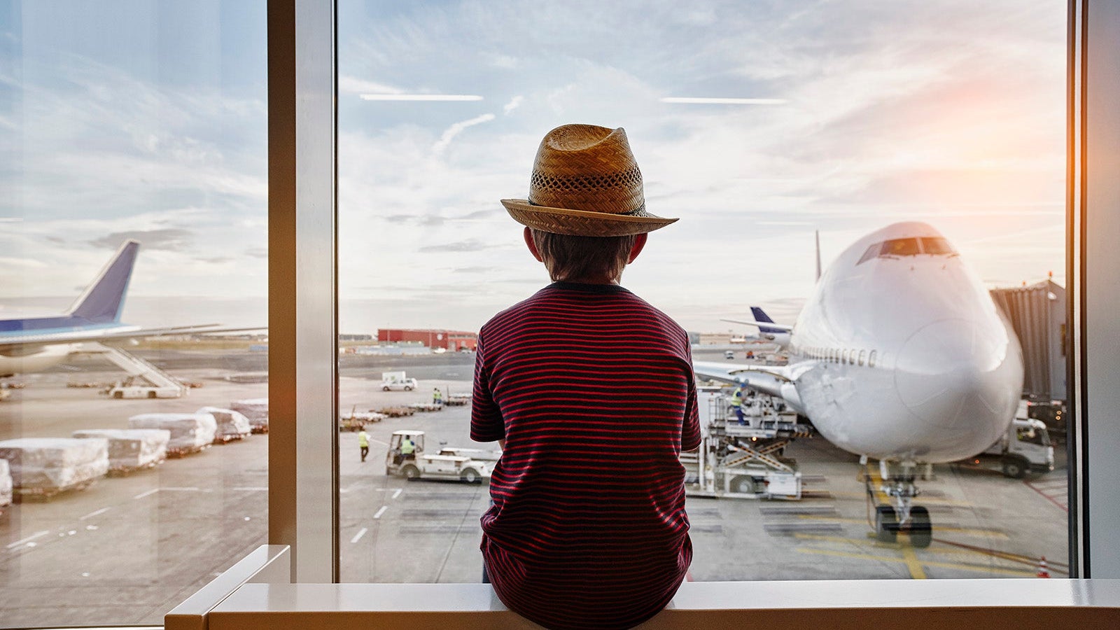 Boy wearing straw hat looking through window to airplane on the apron
