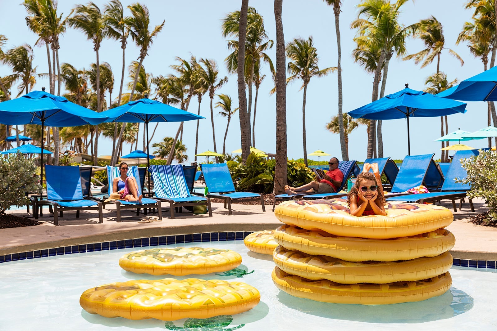 a child lays on a stack of pool floats while adults rest on lounge chairs near the pool
