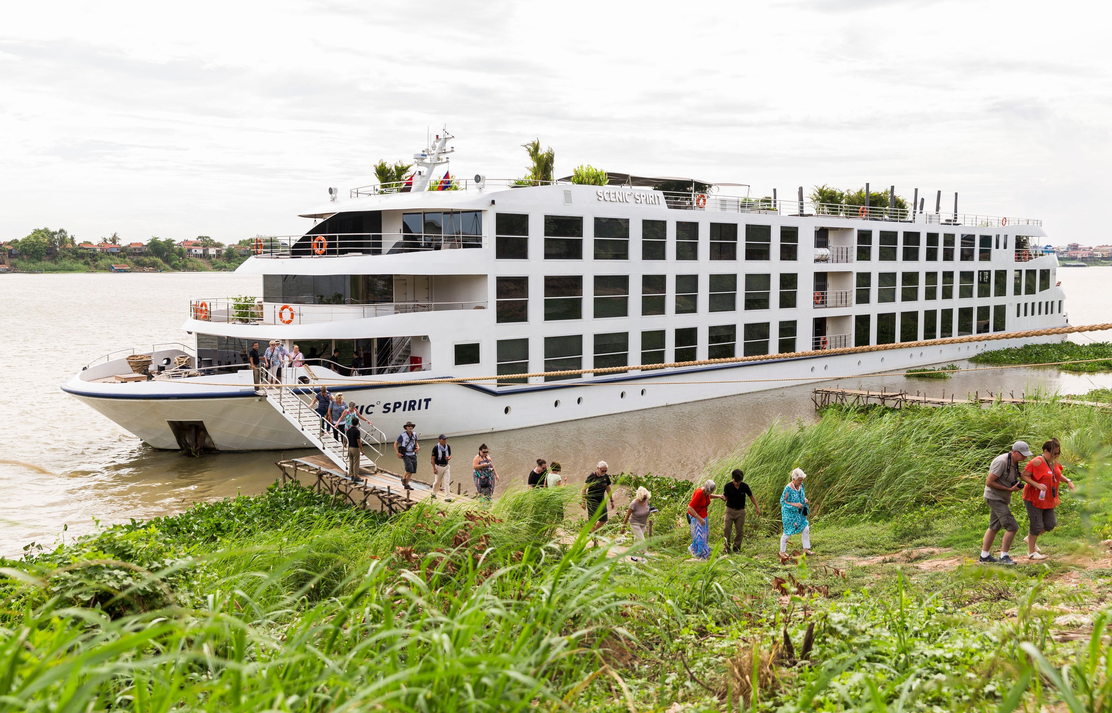 A riverboat docked along a grassy riverbank in the muddy Mekong with passengers boarding via a rope bridge