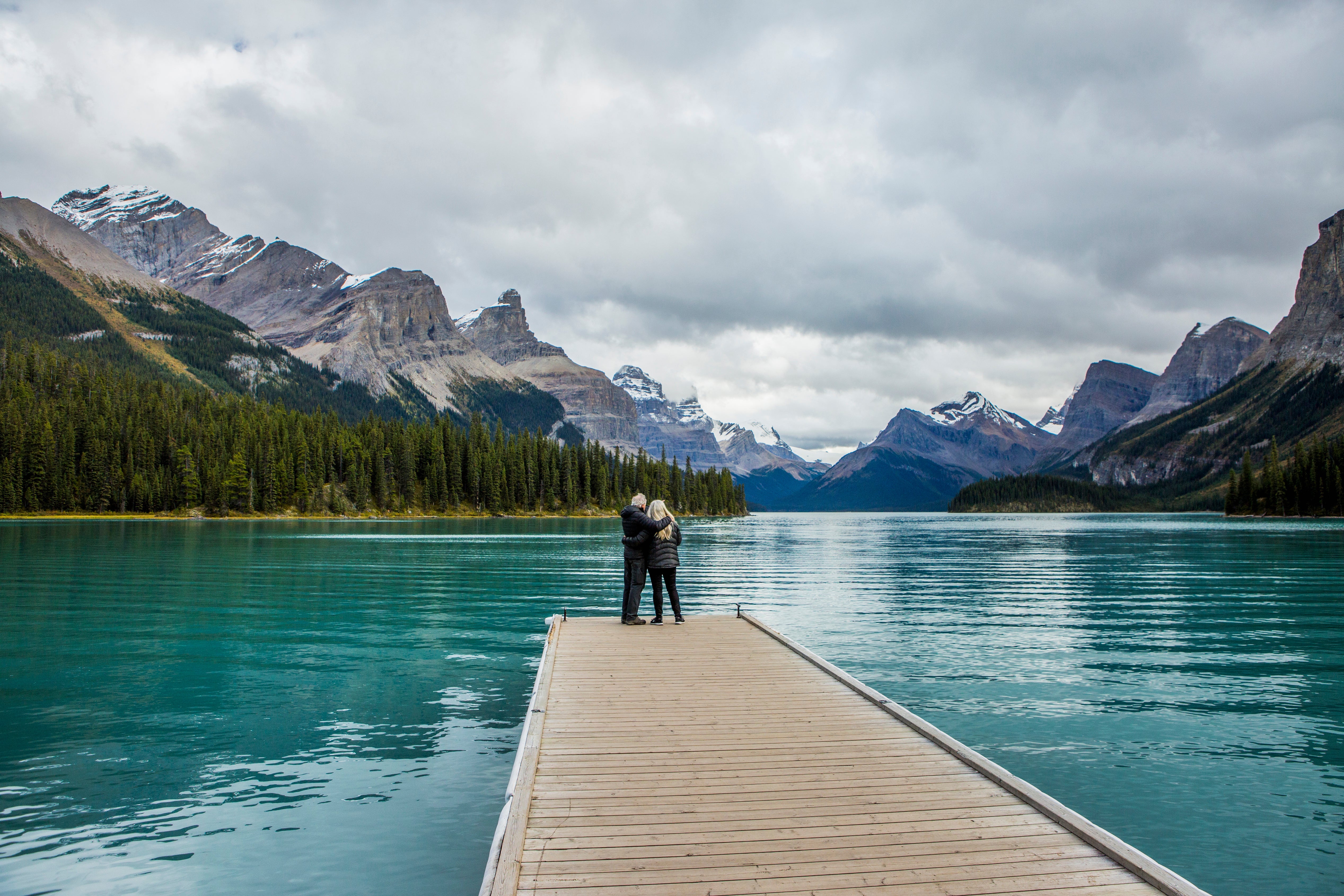 A couple stands on a small jetty over a mountain lake