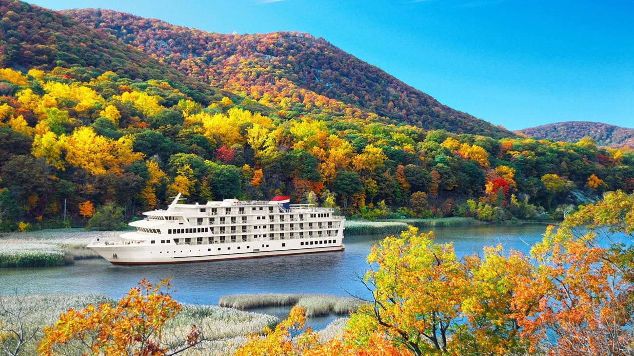 A white riverboat sailing between moutains bright with fall foliage