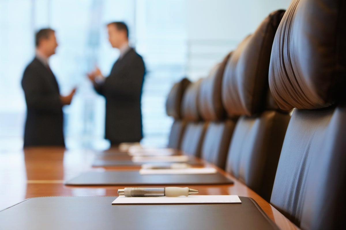Conference room table lined with leather chairs and two men in suits standing in the background