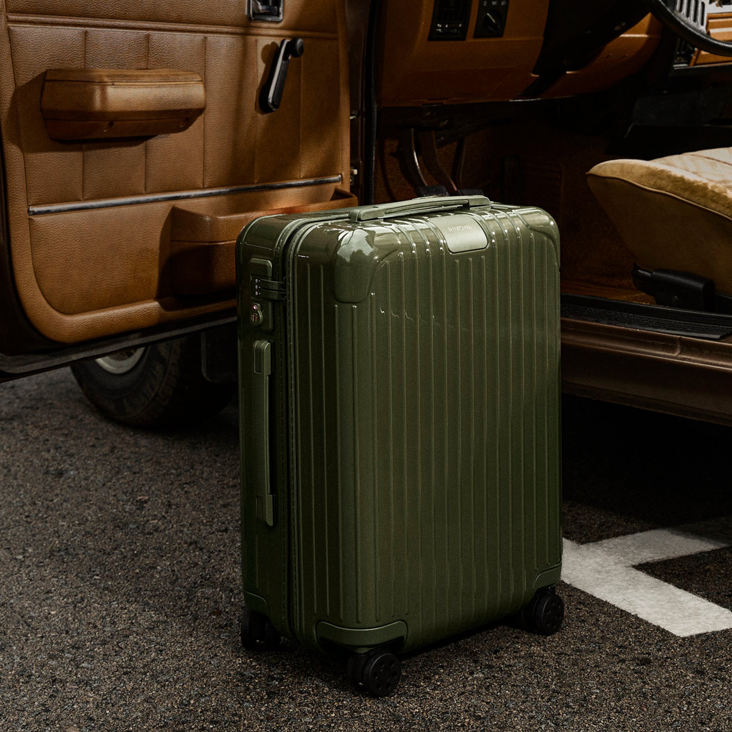 A dark green and glossy RIMOWA carry-on bag sitting on the ground next to an open car door.