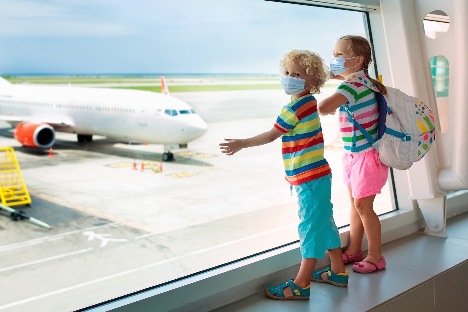 Two children wearing masks looking out the window at an airplane.