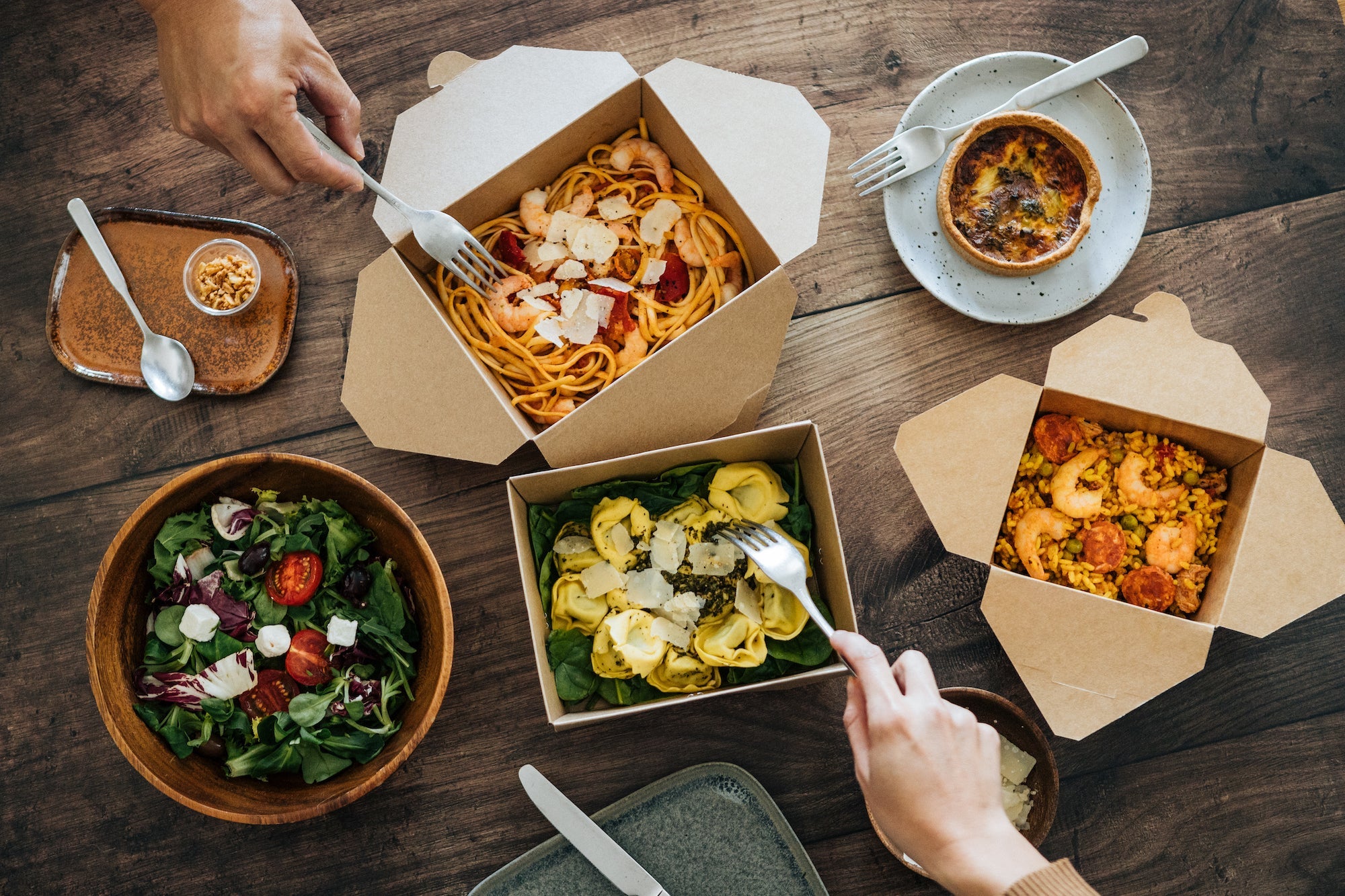 Flat lay view of couple eating delicious takeaway meals on dinner table - with pasta, vegetable salad and seafood risotto.