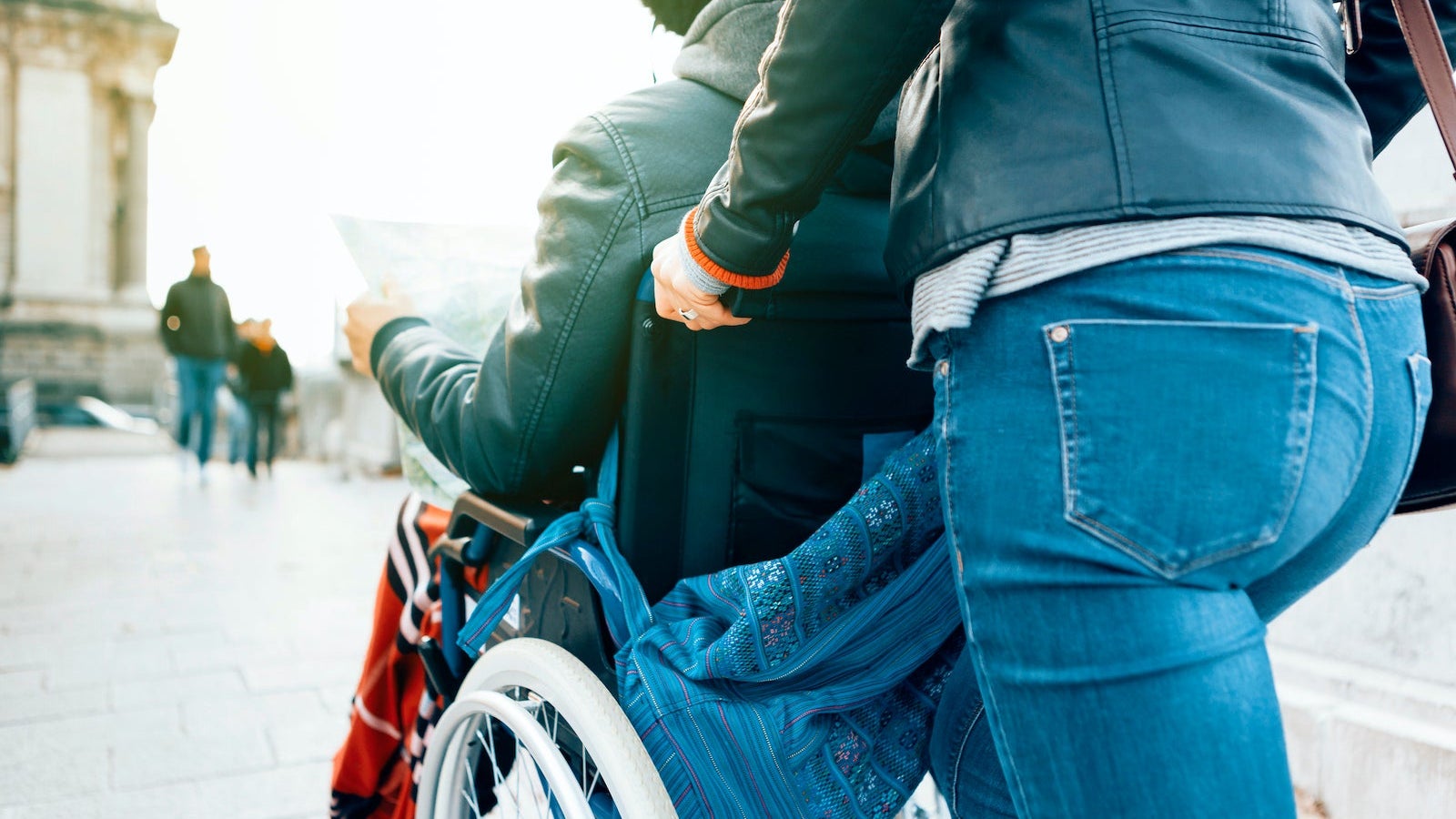 Side View Of A Man Sitting In A Wheelchair While A Female Person Is Pushing Him Around The City