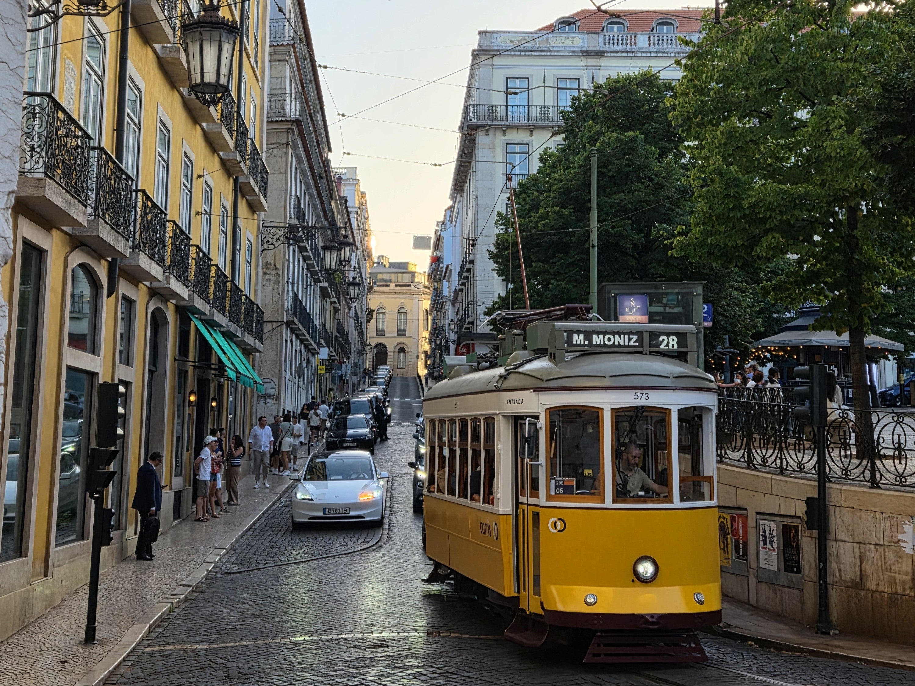yellow tram driving down street between buildings with cars behind