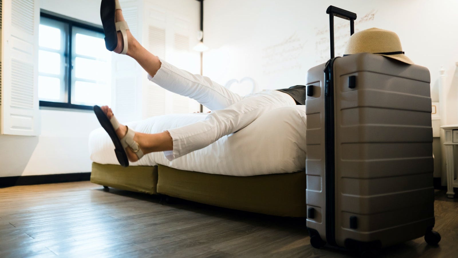 Young female traveller relaxing on bed in a hotel room, with a suitcase and straw hat by the bed