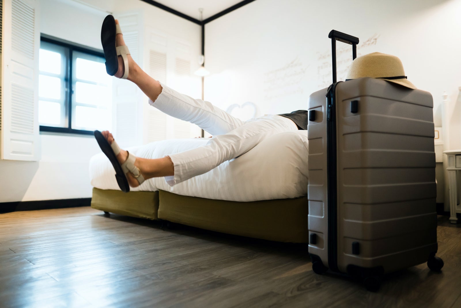 A woman kicks her legs up and she falls onto her hotel bed. Suitcase and hat next to her on the right.