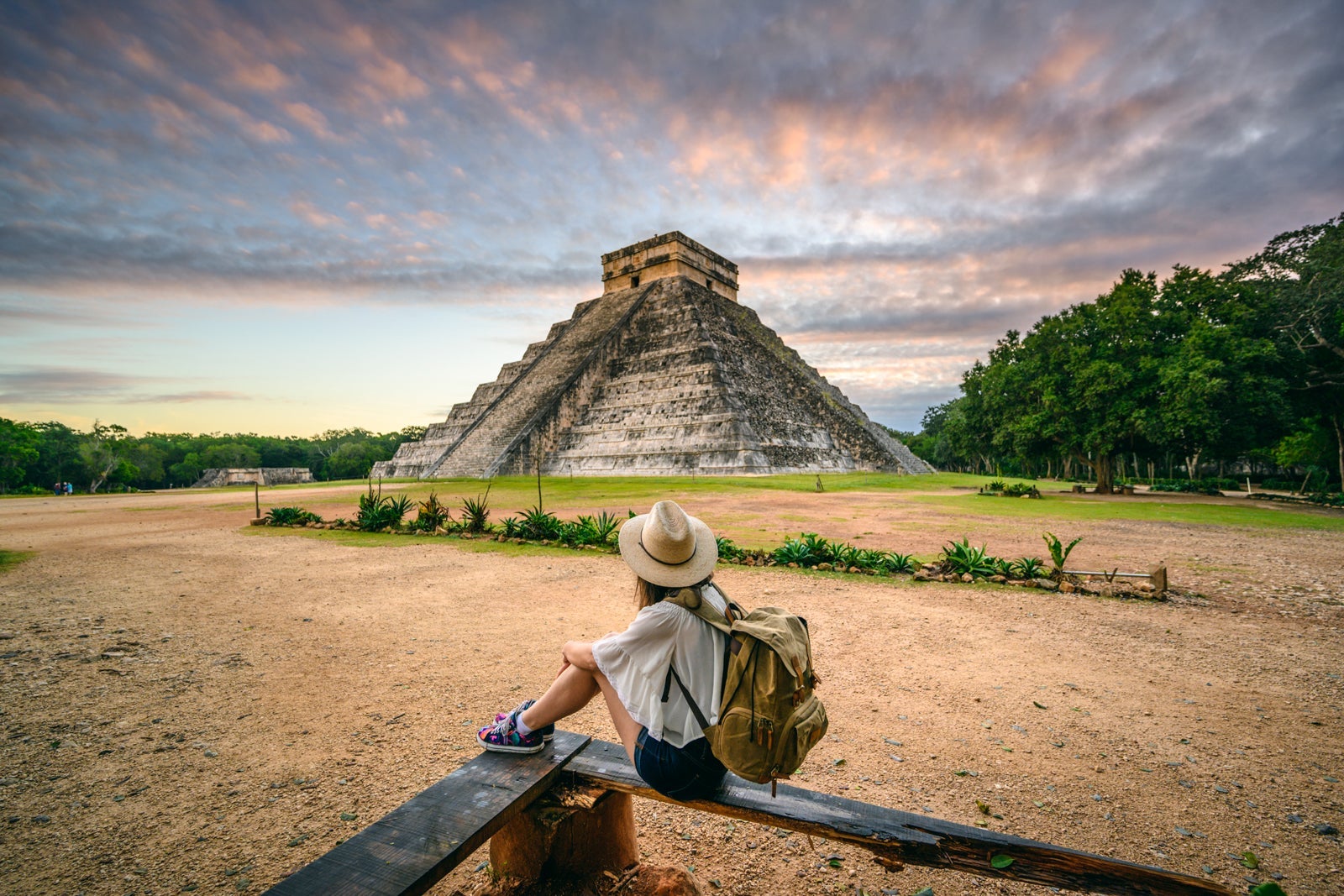 A woman sit in the foreground while Chichen-Itza looms in the background.