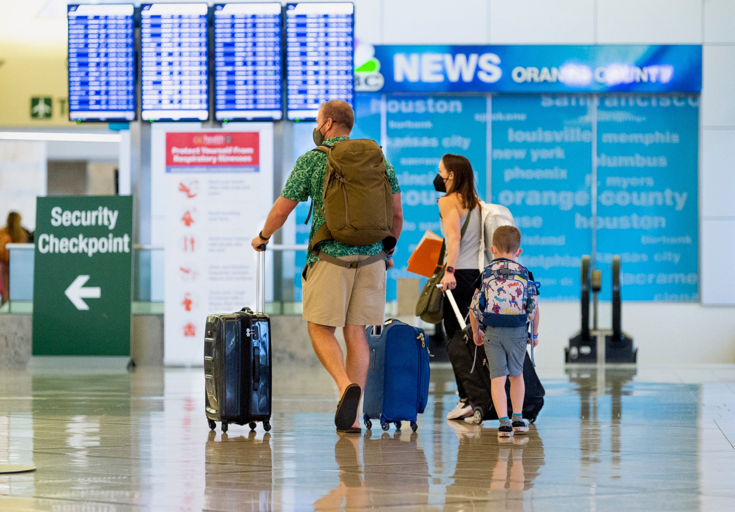 A man, woman, and young child, all wearing masks, wheel luggage through an airport.