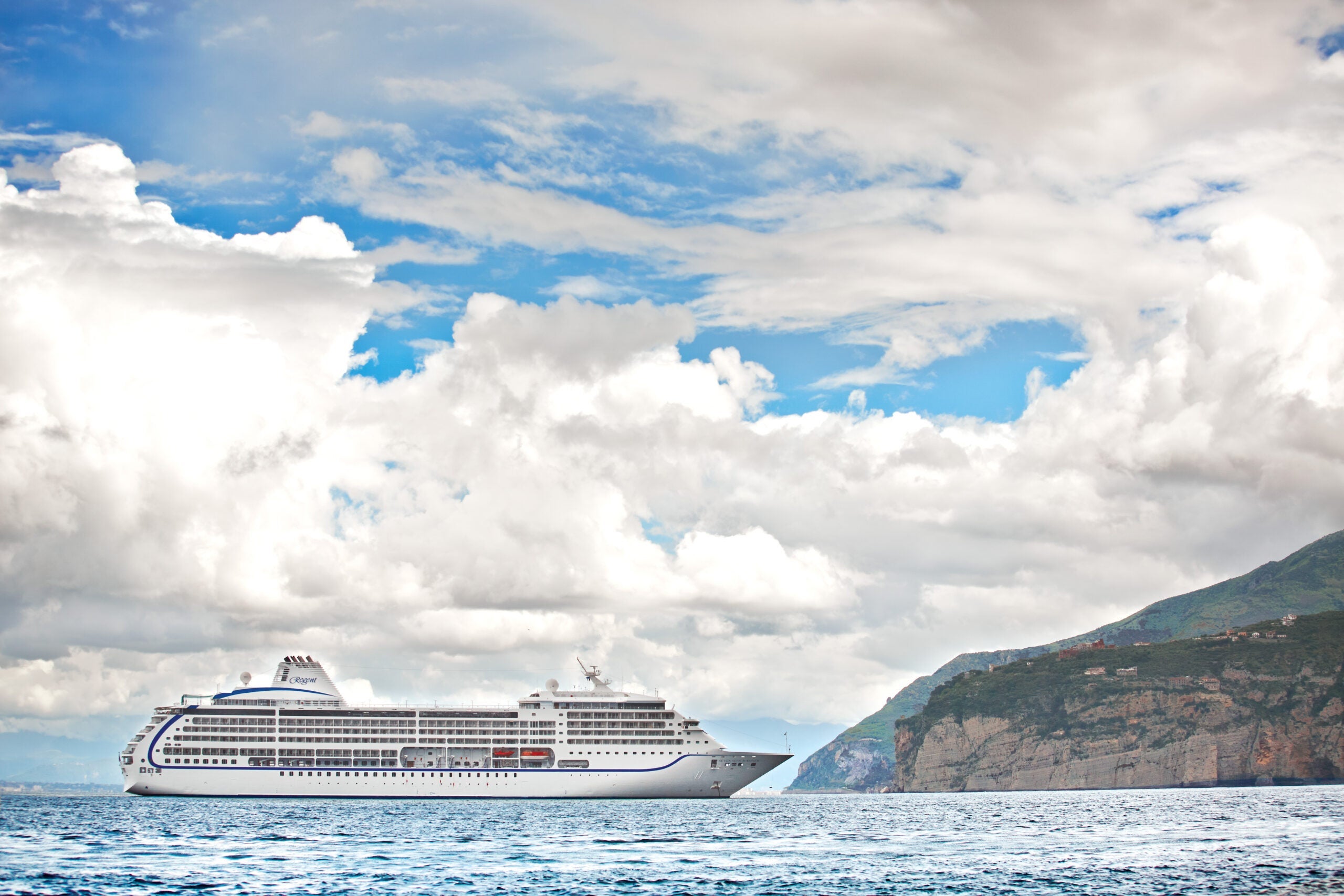 A cruise ship floats on wavy blue water near a rock outcropping with lots of fluffy, white clouds in the blue sky background