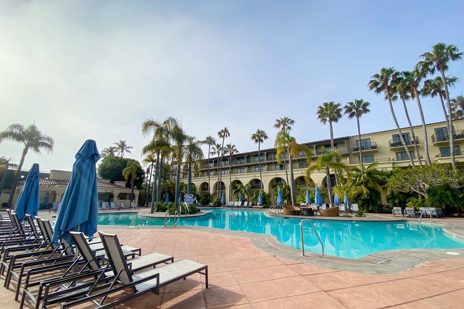 Hotel pool on a sunny day, surrounded by loungers with palm trees and building in the background