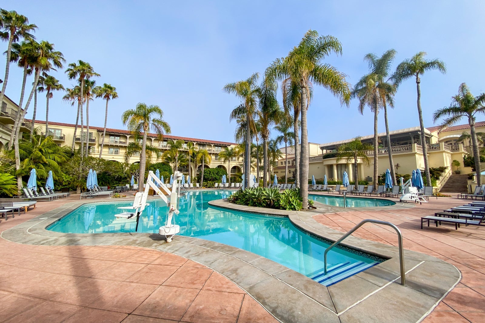 Hotel pool on a sunny day, surrounded by loungers with palm trees and building in the background