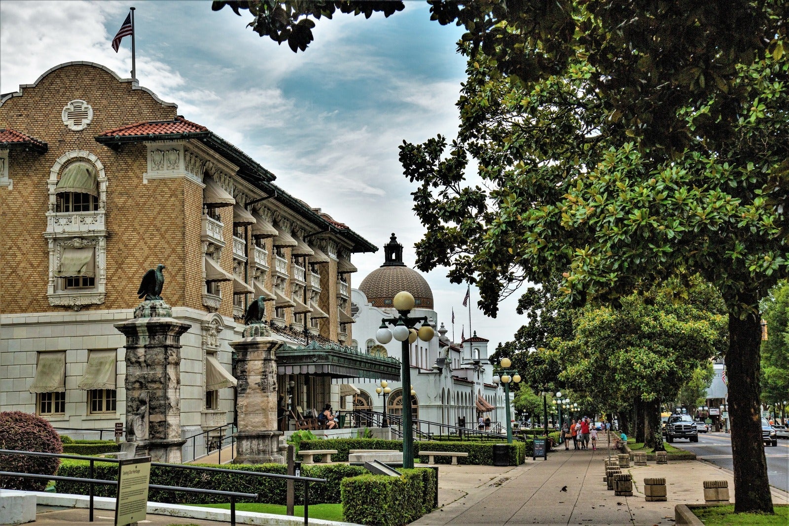 Two large buildings sit prominently along a sidewalk. Two stone pillars with eagles atop them mark an entrance to the park on the left, while a row of Magnolia trees provides a barrier to the street.