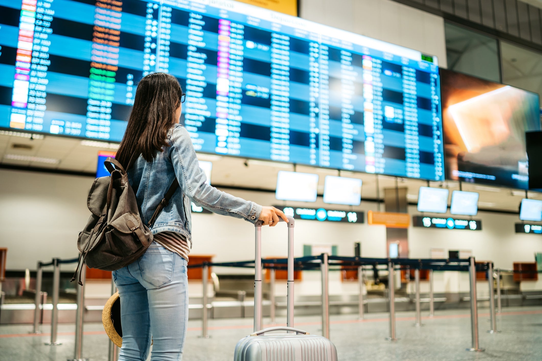 A woman with a backback and a suitcase looks at a large display board in an airport
