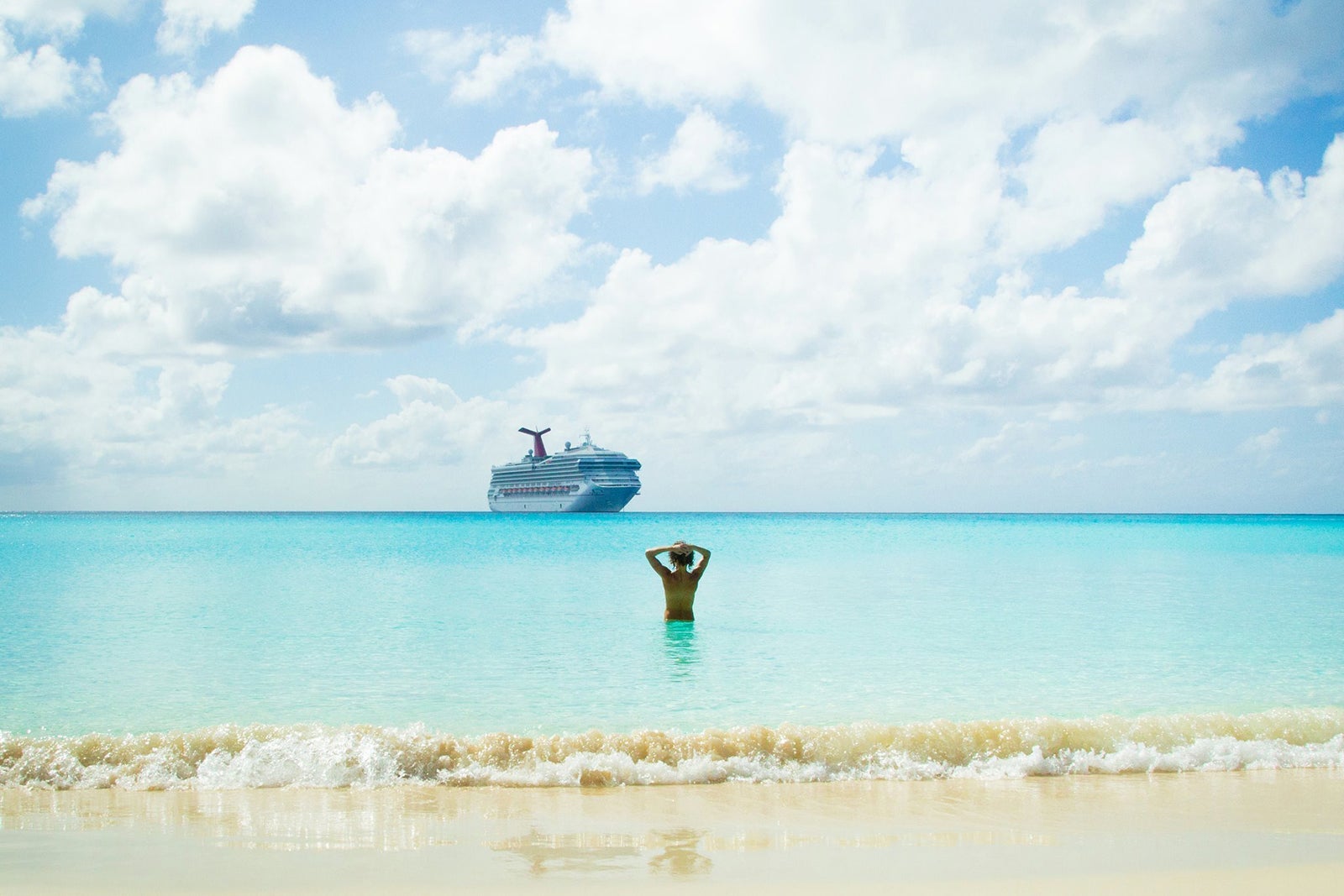 A shirtless person standing in waist-deep light-blue water with a cruise ship and blue sky with white clouds in the background and sand in the foreground