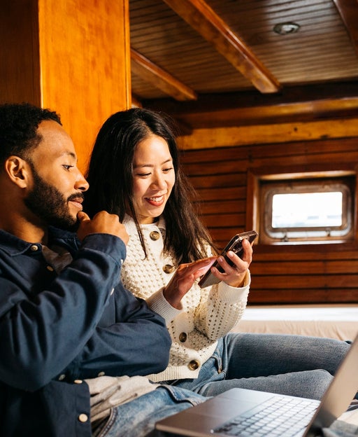 Young mixed race couple using on a laptop sitting on a bed in a cabin of a vintage sailing boat