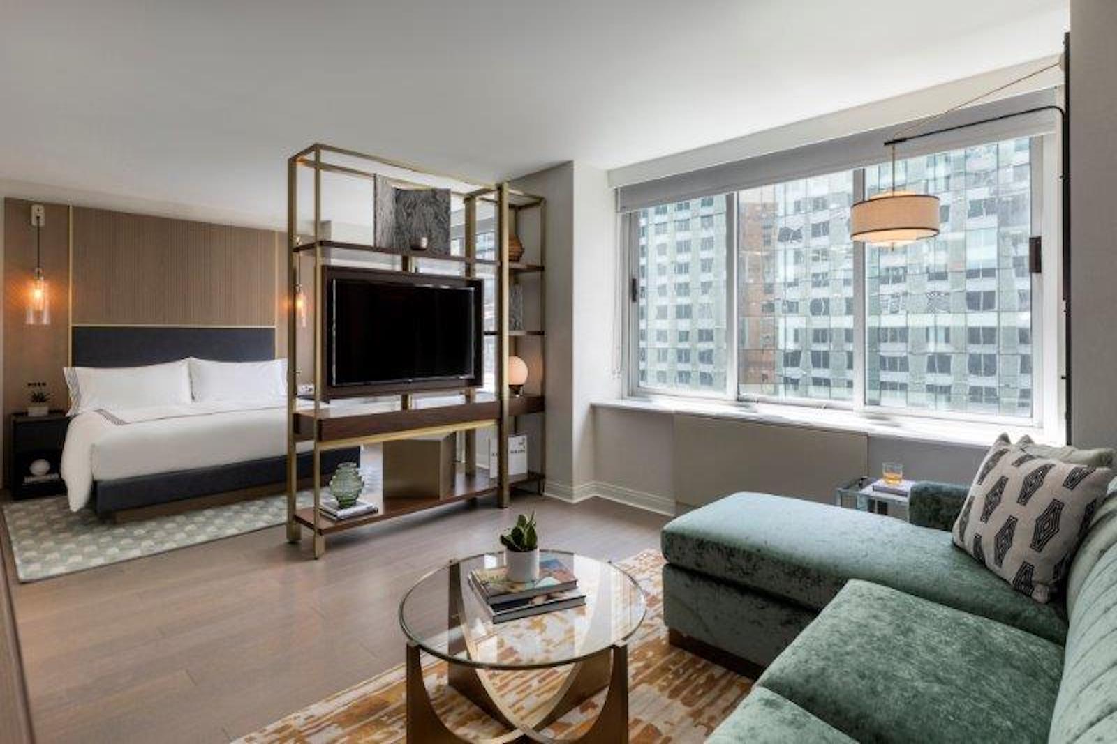 large hotel room with green couch, tv on bookshelf, bed, and views of New York City