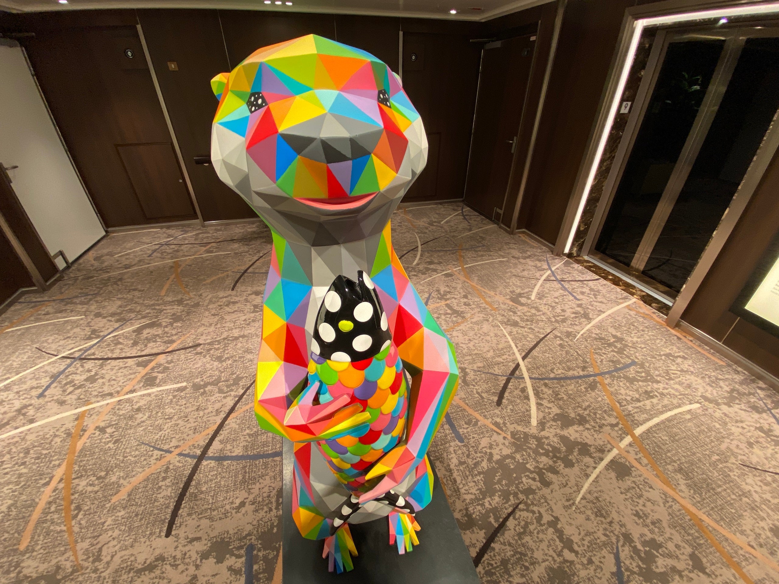 A colorful otter sculpture holding a polka-dotted fish stands in an elevator bay on a Holland America cruise ship.