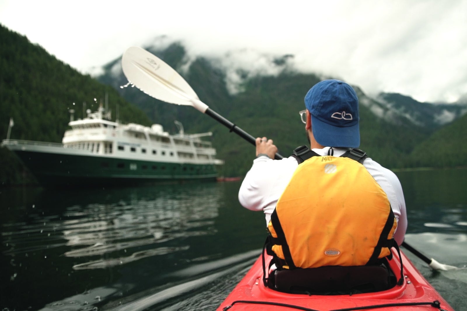 Man in baseball hat kayaking in Alaska's Misty Fjords near expedition cruise ship