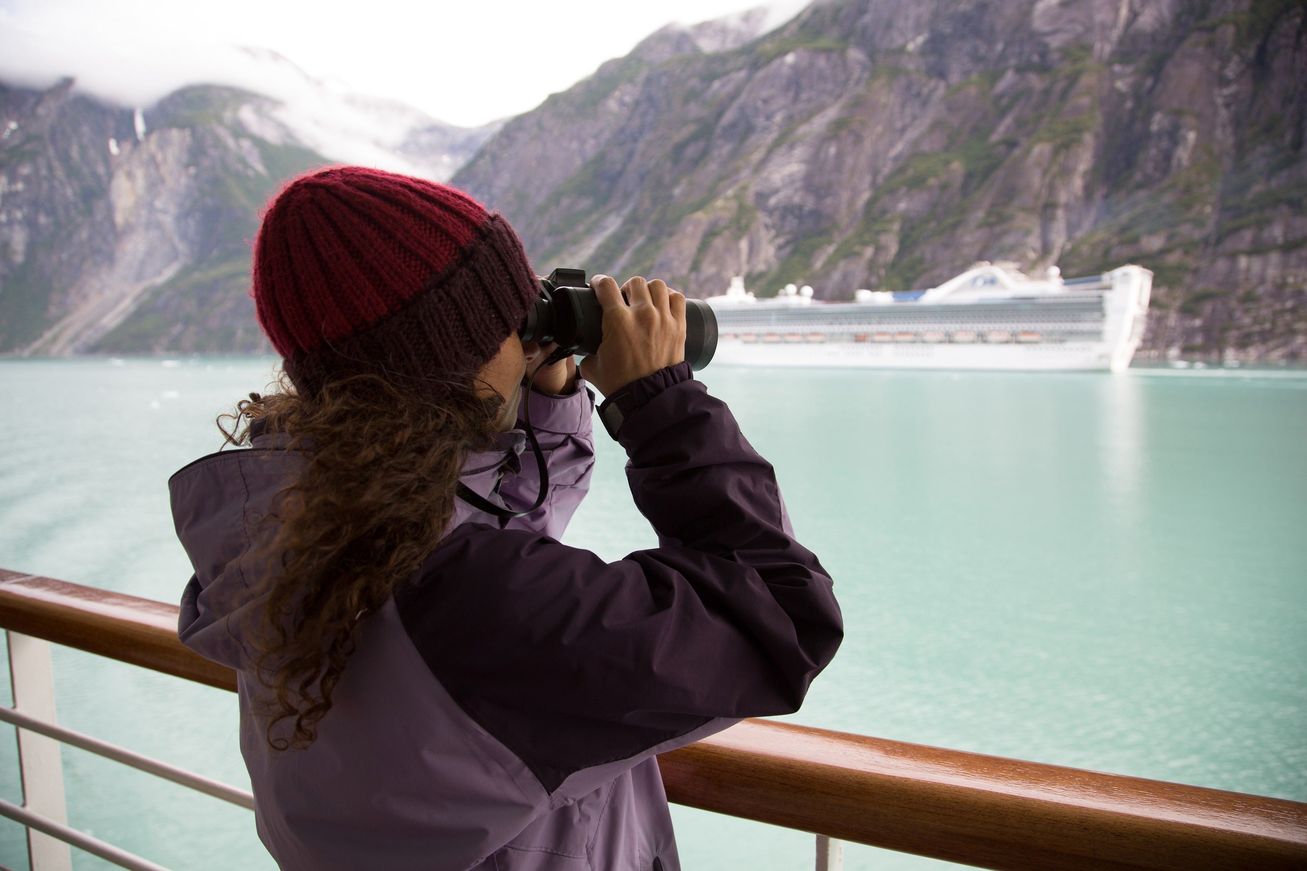 Woman in a coat and hat against a cruise railing with binoculars looking at a glacier in Alaska