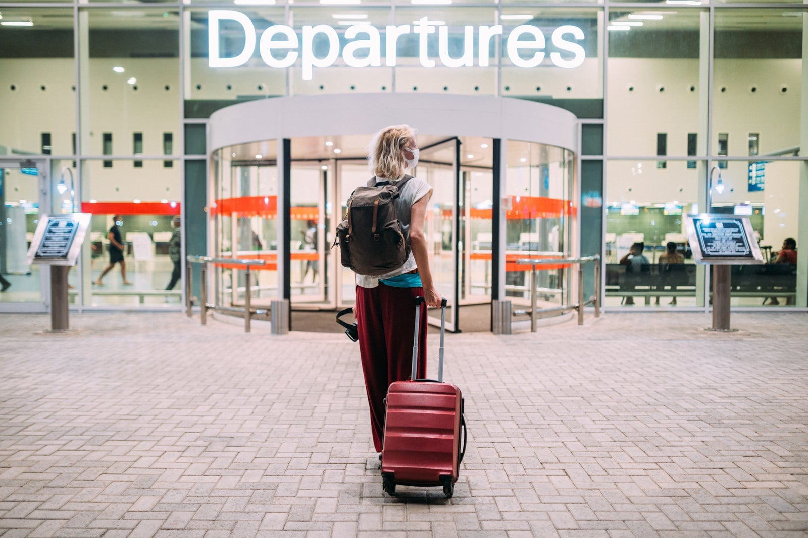 Woman with red rollaboard walking toward revolving doors under sign reading Departures