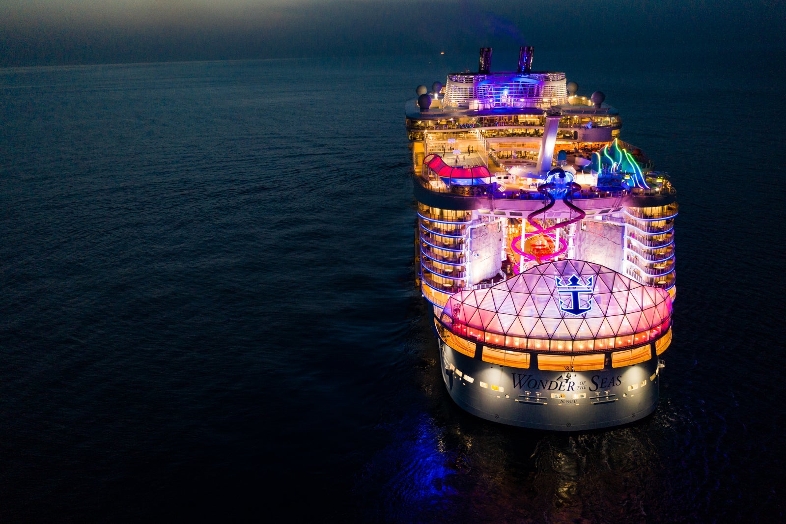 Aerial image of Royal Caribbean's Wonder of the Seas lit up with colored lights surrounded by the dark ocean at nighttime
