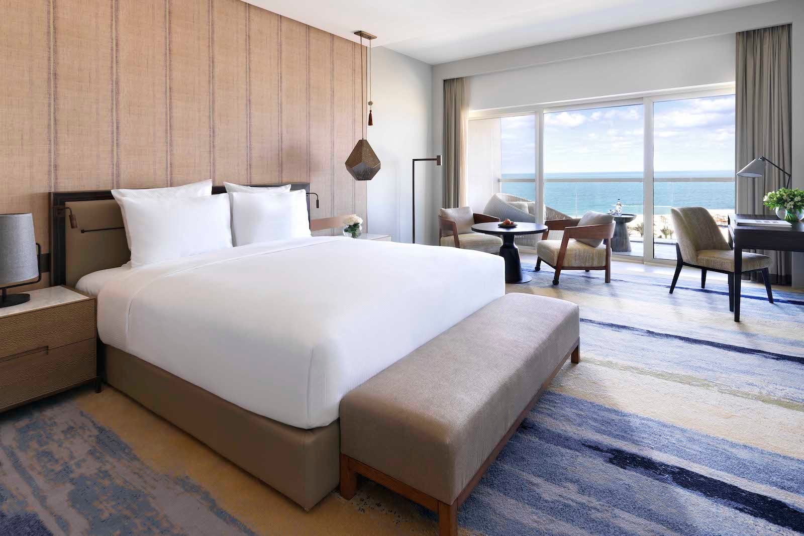 hotel room with views of the ocean, table with chairs, desk and chair and large bed with white linens against wooden wall