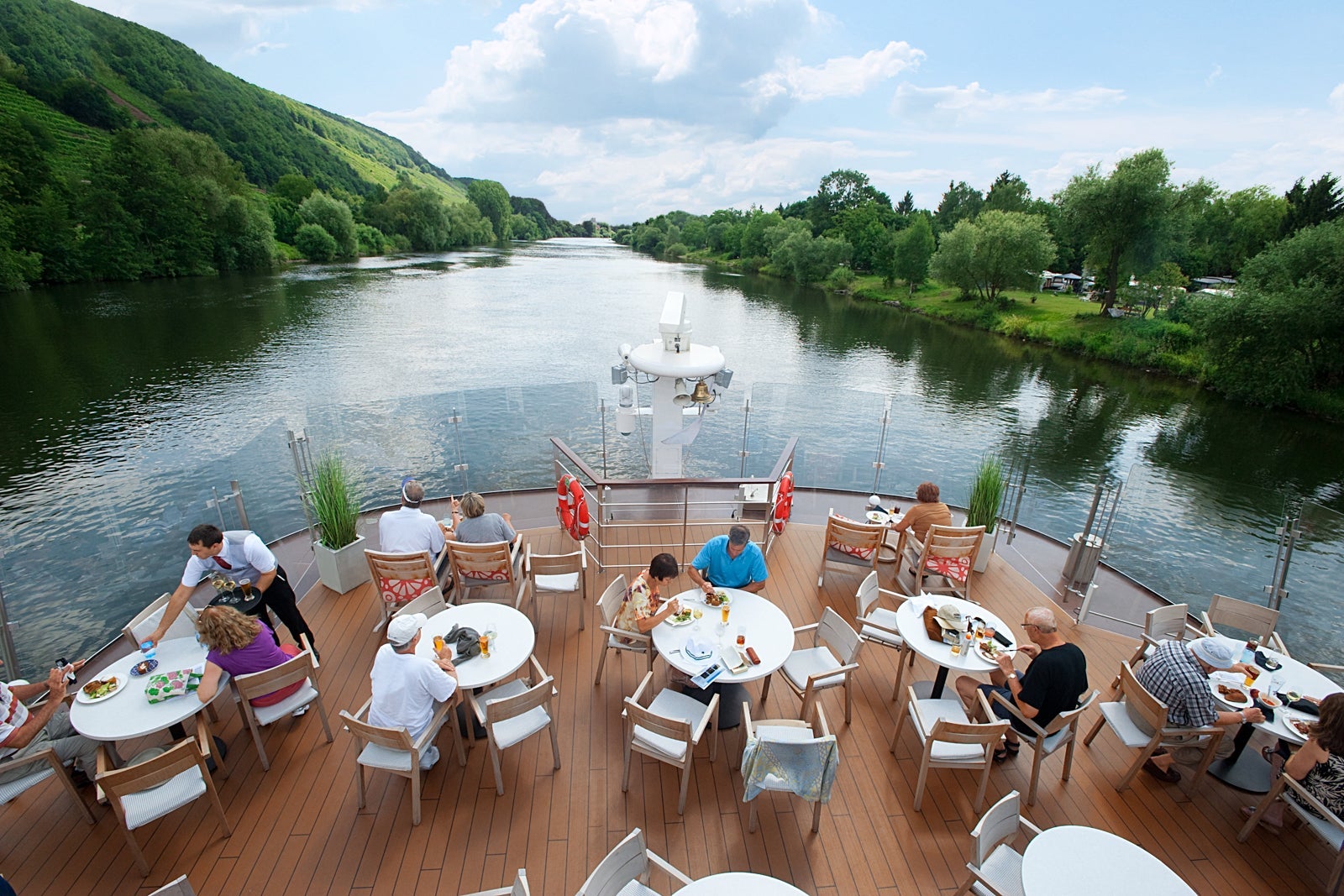 An outdoor terrace on a cruise ship facing the wake with people dining at several round tables with chairs