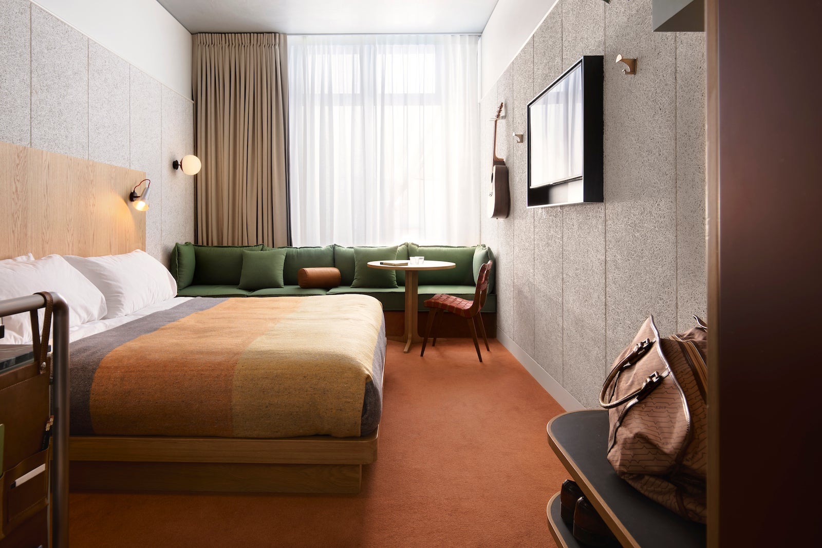 rendering of trendy hotel room with bed, red-orange carpet, green couch and TV on wall
