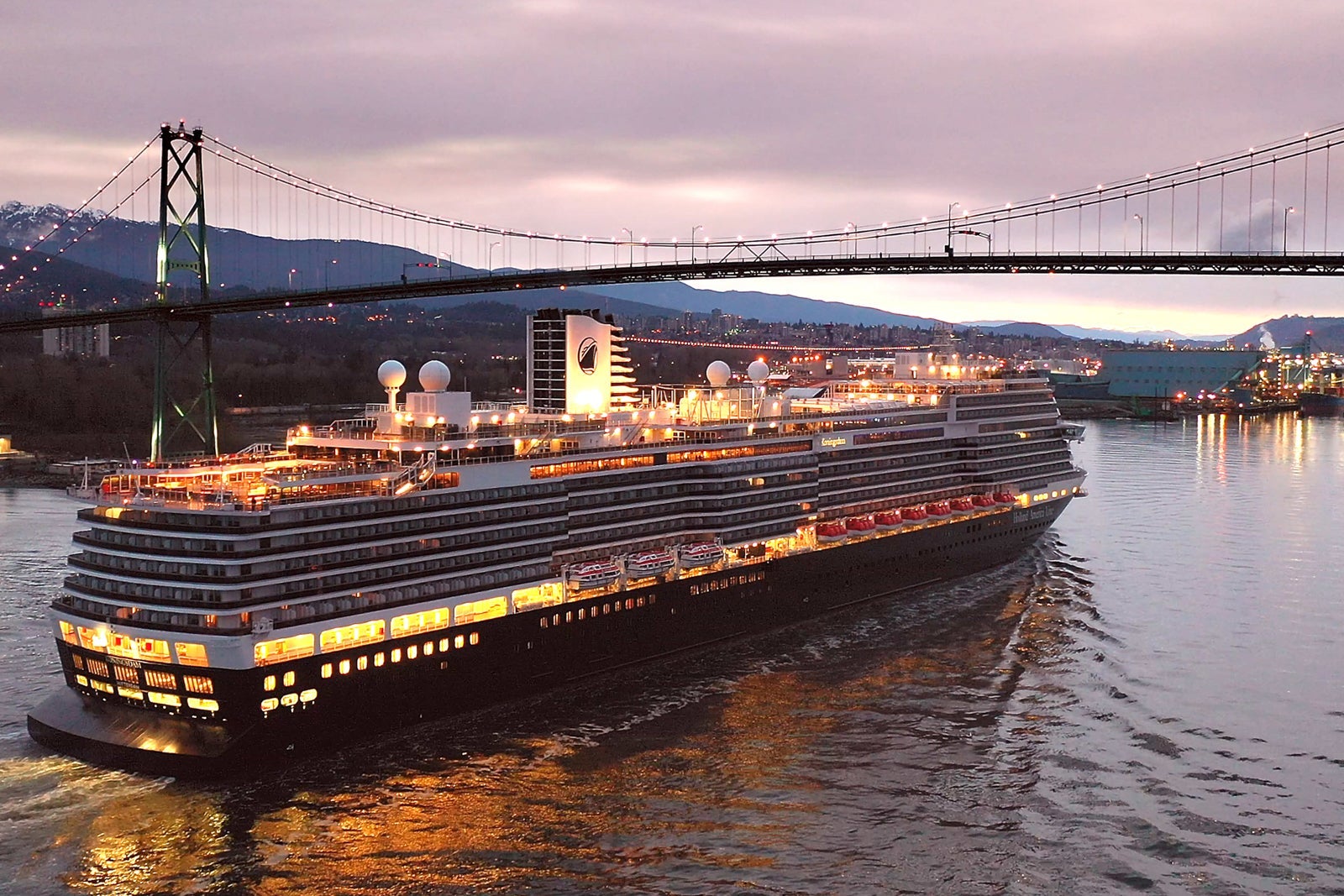 A cruise ship sails along the coast and prepares to pass under a bidge near dark with its lights aglow