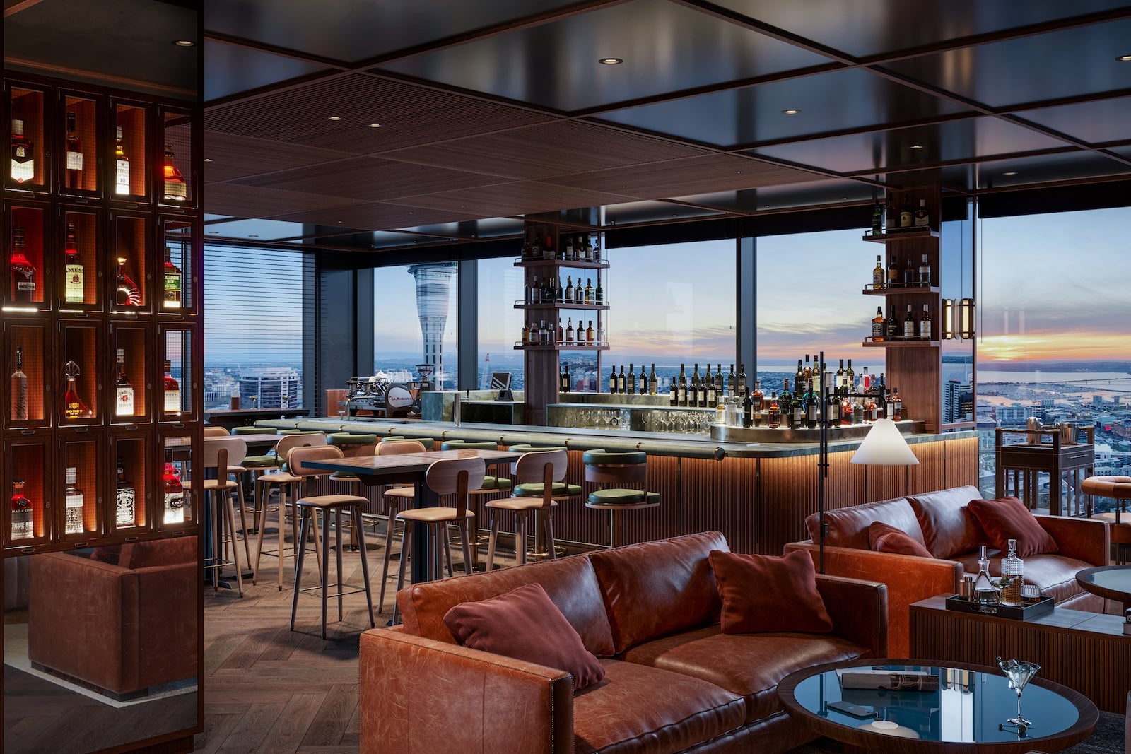 large and airy hotel bar with leather seats, glass shelves with bottles and views of a sunset