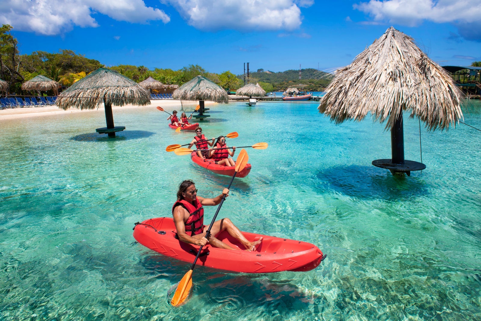 People in red kayaks with orange paddles on turquoise water among tiki huts