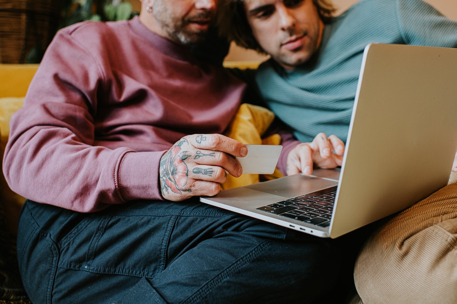 A gay couple sit on a yellow sofa and use a laptop together. One man holds a credit card. They are relaxed and content as they look at the screen.
