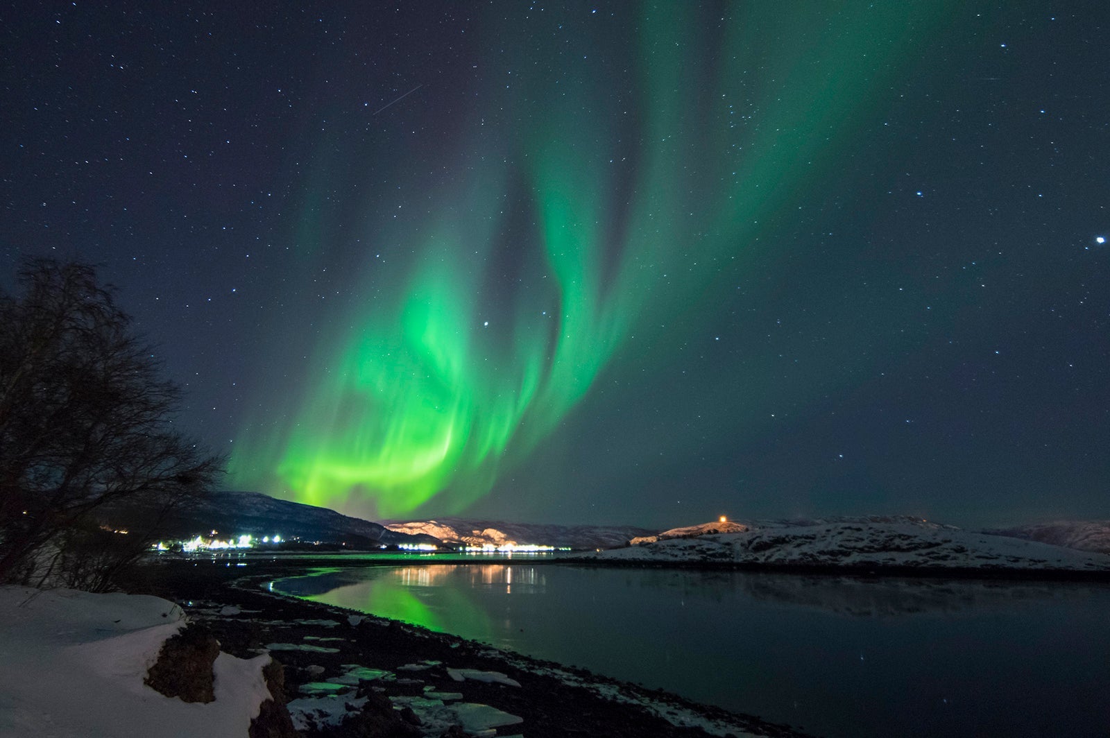 Northern lights over brightly lit homes and a peaceful lake in Norway