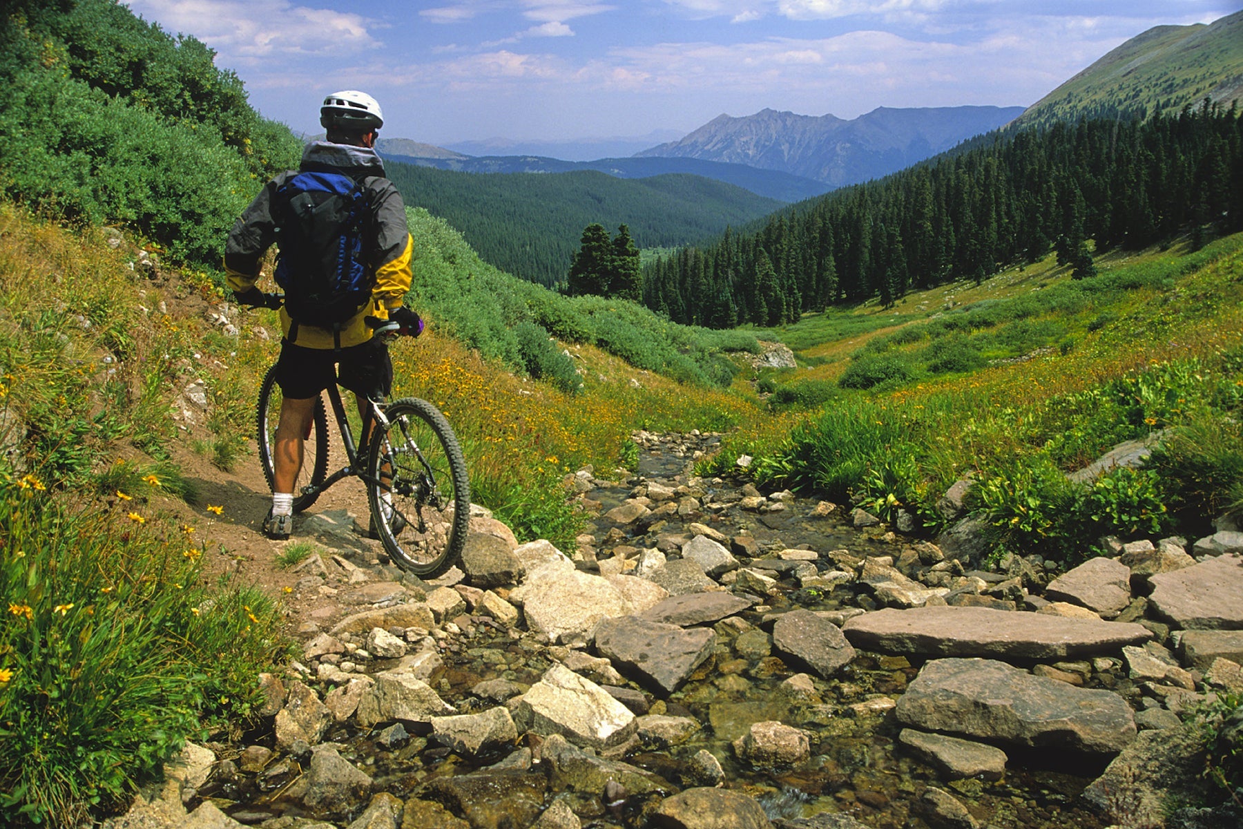A man in a yellow and black windbreaker jacket with a black backpack sits on a bike overlooking a mountain and field.