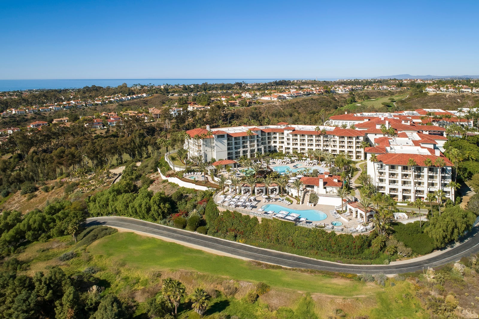 aerial view of large white resort with red roofs, pool and ocean in distance