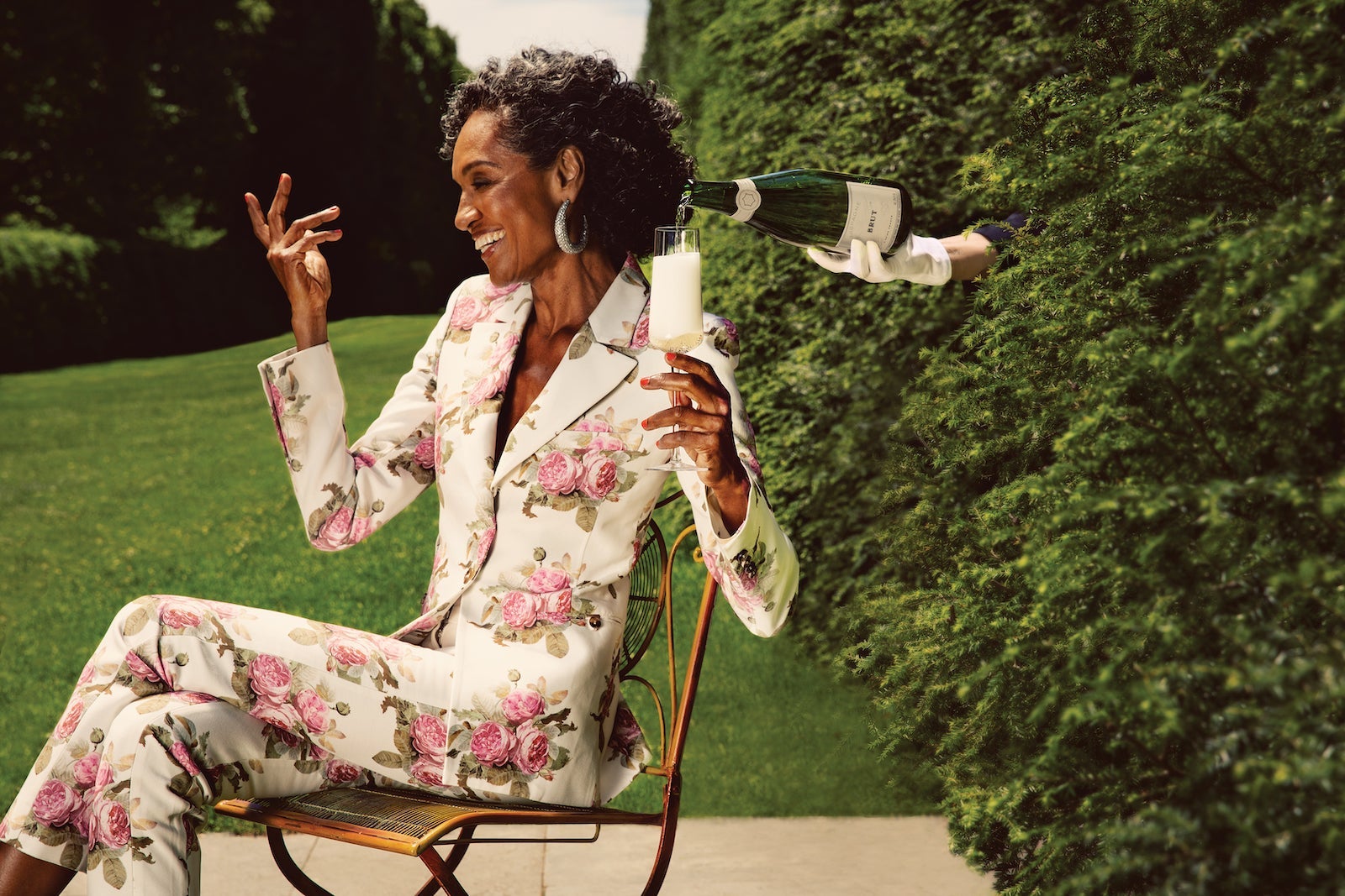 woman in floral suite sitting in chair holding glass of champagne, a hand reaches through bushes to pour champagne