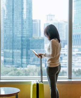 woman on phone with suitcase standing in front of window with city skyline in background