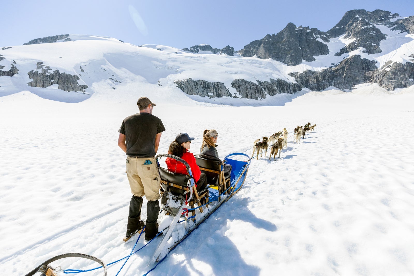 A team of several dogs pulls a two-person sled through the snow with a musher standing behind
