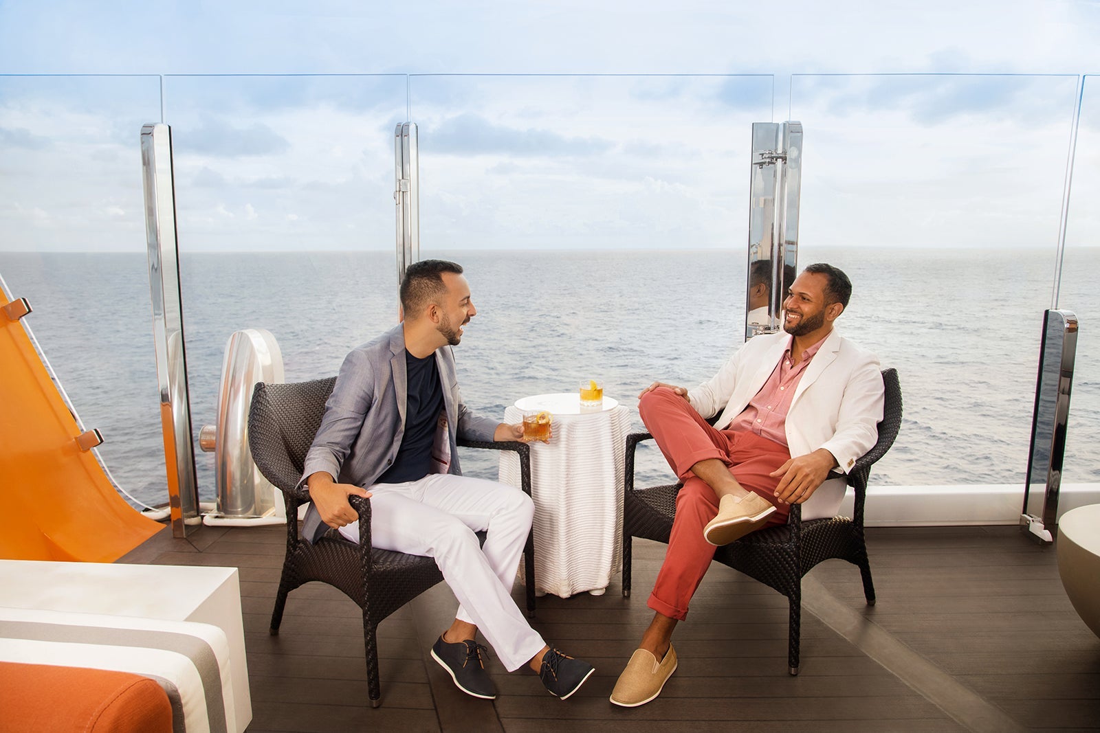 Two passengers sit at a table and enjoy cocktails outdoors on a cruise ship