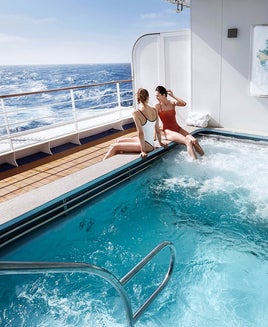 Two women in bathing suits sit on the edge of a large spa hot tub on an outer deck of a cruise ship overlooking the water