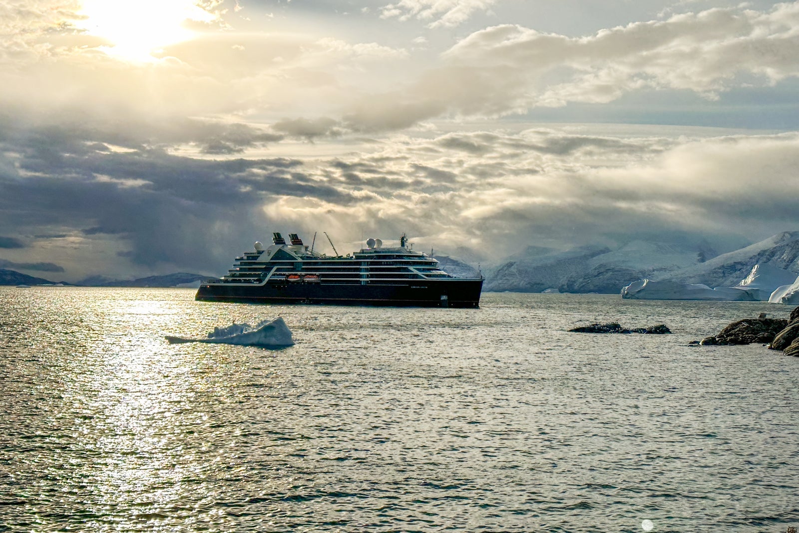 Seabourn Venture anchored with sun breaking through the clouds behind it and icebergs floating in front