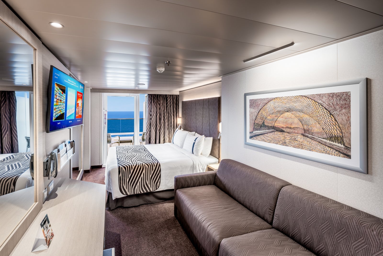 A cruise ship cabin with a sofa, queen bed, TV and balcony